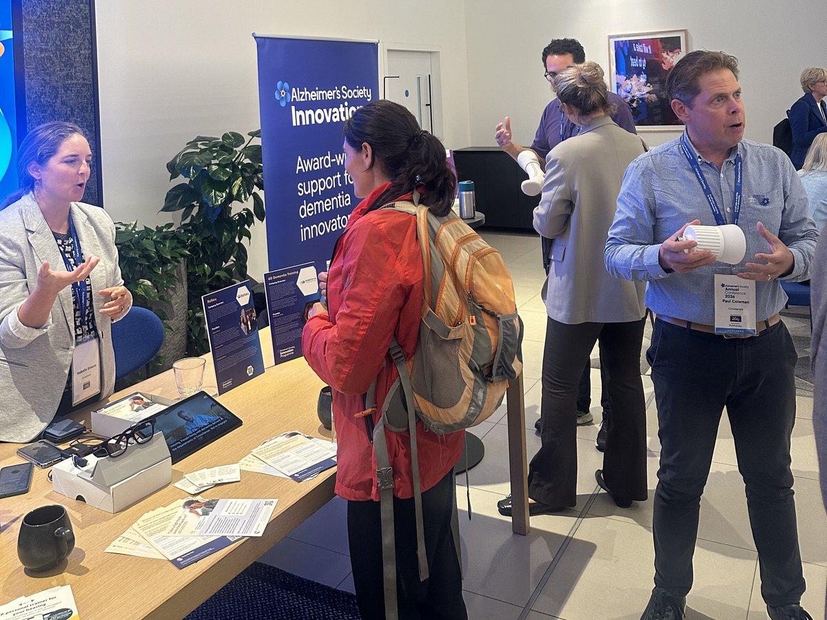 A CrossSense co-founder talks with visitors at a stand with smart glasses and a tablet, beside an Alzheimer's Society Innovation banner.